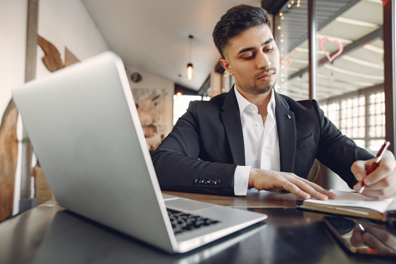 A businessman taking notes beside a laptop in a cafe